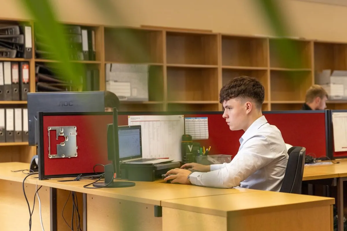 Un impiegato al computer in un ufficio, con la parete di faldoni alle spalle